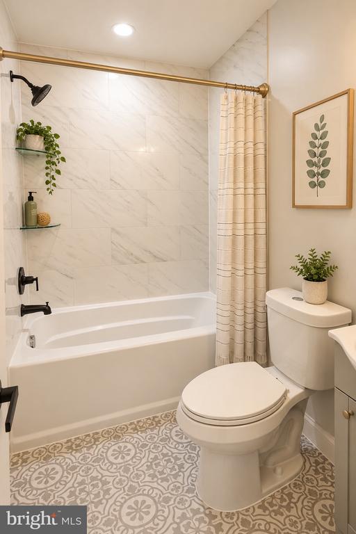 Bathroom with a white tub, marble-look tile, brass shower rod, and patterned gray floor.