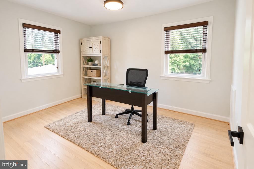 Home office with a dark desk, mesh chair, pale wood shelving, and two windows.