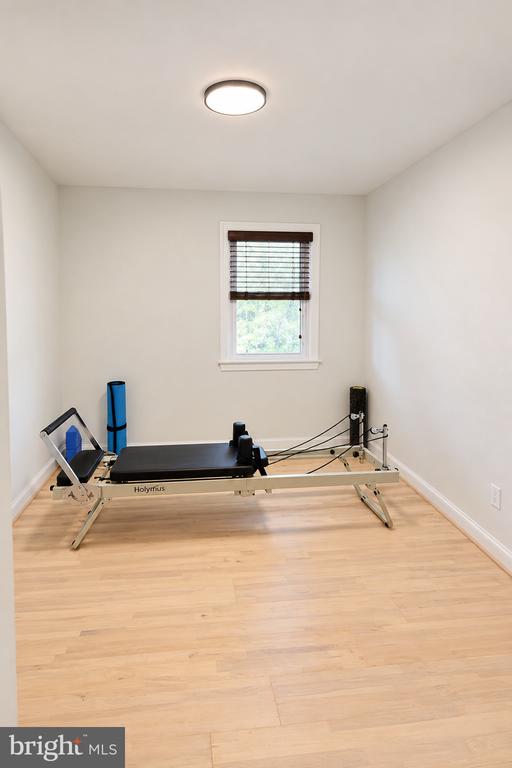 Exercise room with a Pilates reformer, pale wood flooring, and a window with dark blinds.