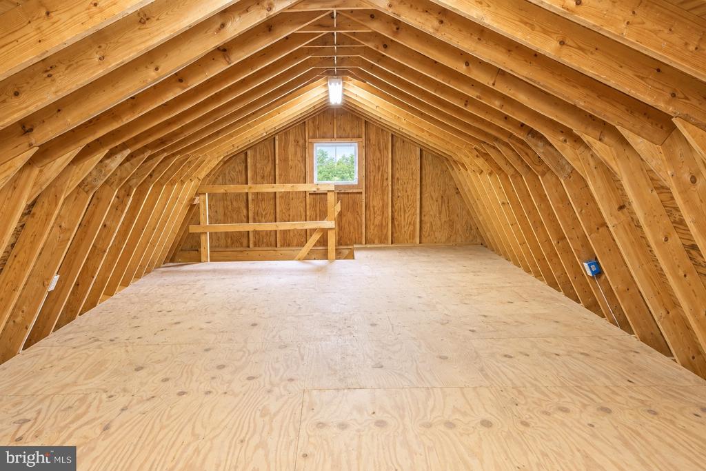 Unfinished attic with exposed wood rafters, plywood flooring, and a small window.