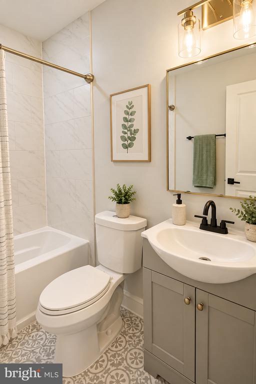 Bathroom with a marble-look tub surround, gray vanity, brass-framed mirror, and patterned floor tile.