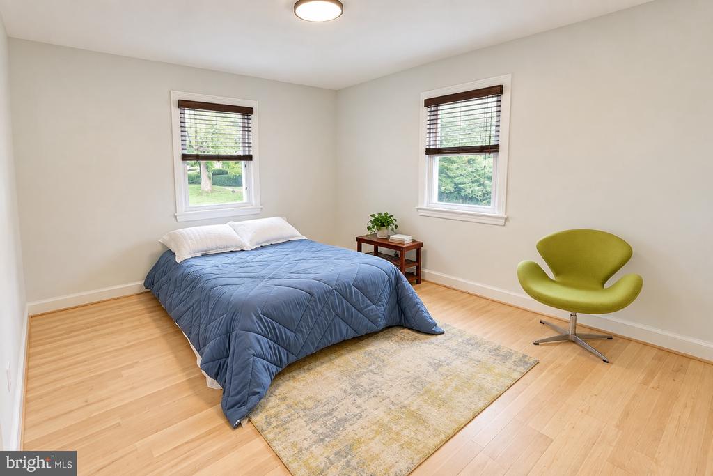 Staged bedroom with a blue bedspread, green accent chair, and two windows with blinds.