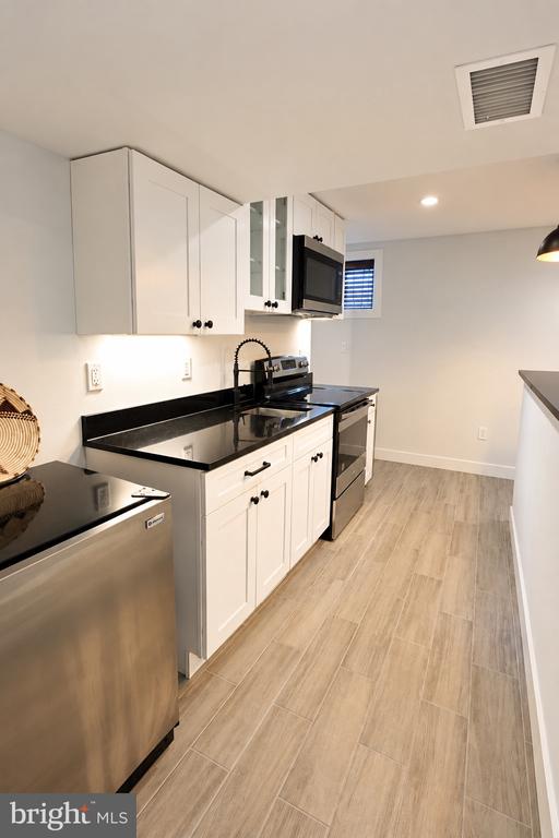 Kitchen with white cabinets, black countertops, stainless steel appliances, and light wood-look tile flooring.