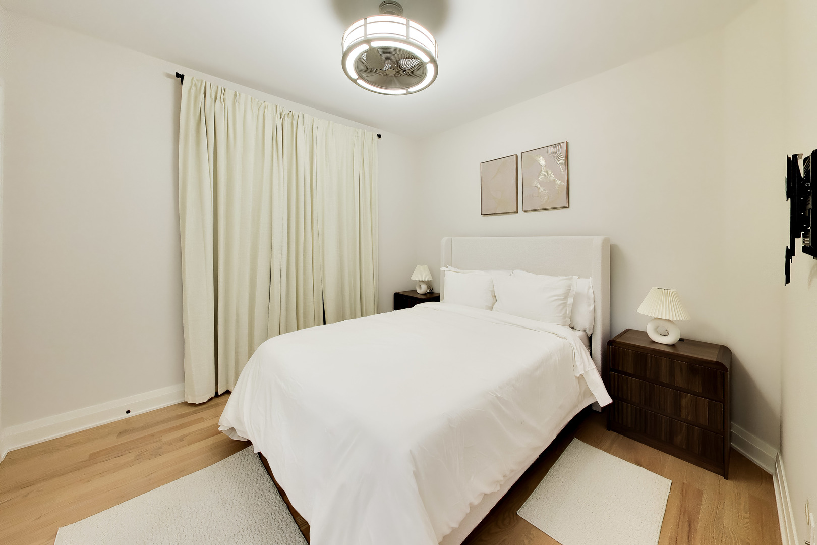 Bedroom with a white upholstered bed, cream curtains, hardwood floors, and dark wood nightstands.