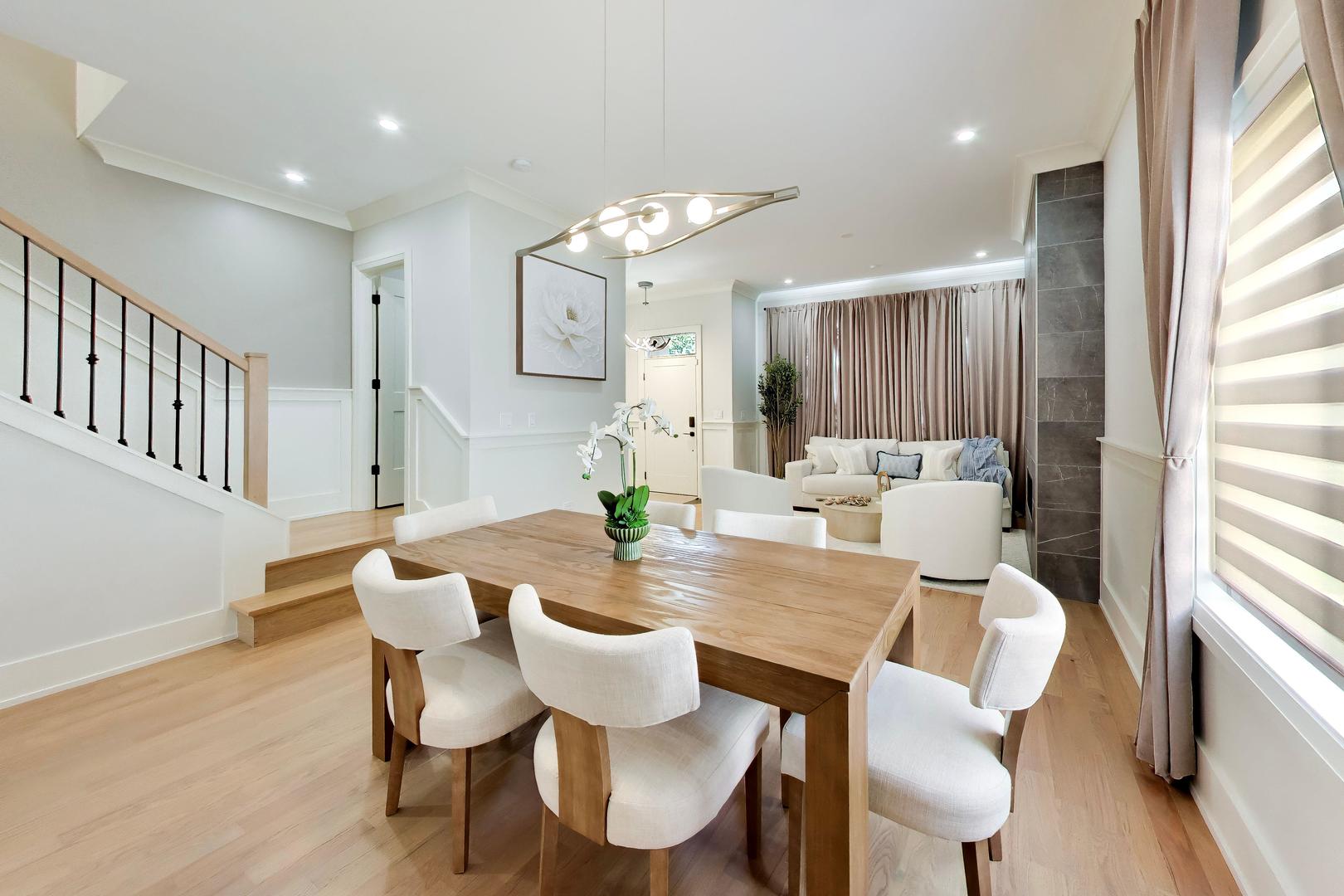 Dining area with a wooden table and white chairs beside a staircase and living room.