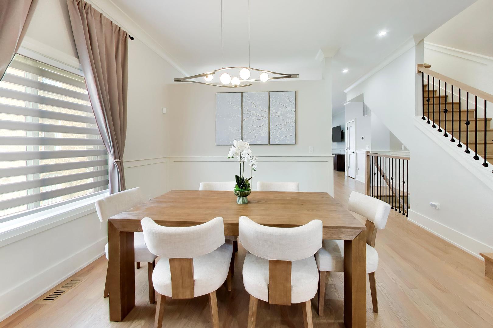 Dining room with a large wooden table, white chairs, and an open staircase.