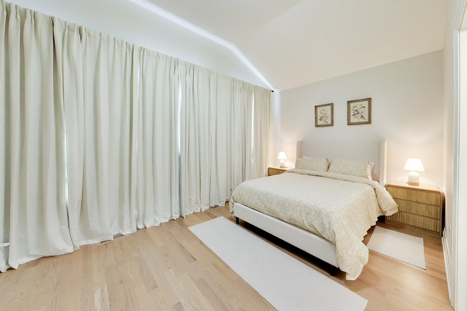 Bright bedroom with a large upholstered bed, pale wood floors, and floor-to-ceiling curtains.