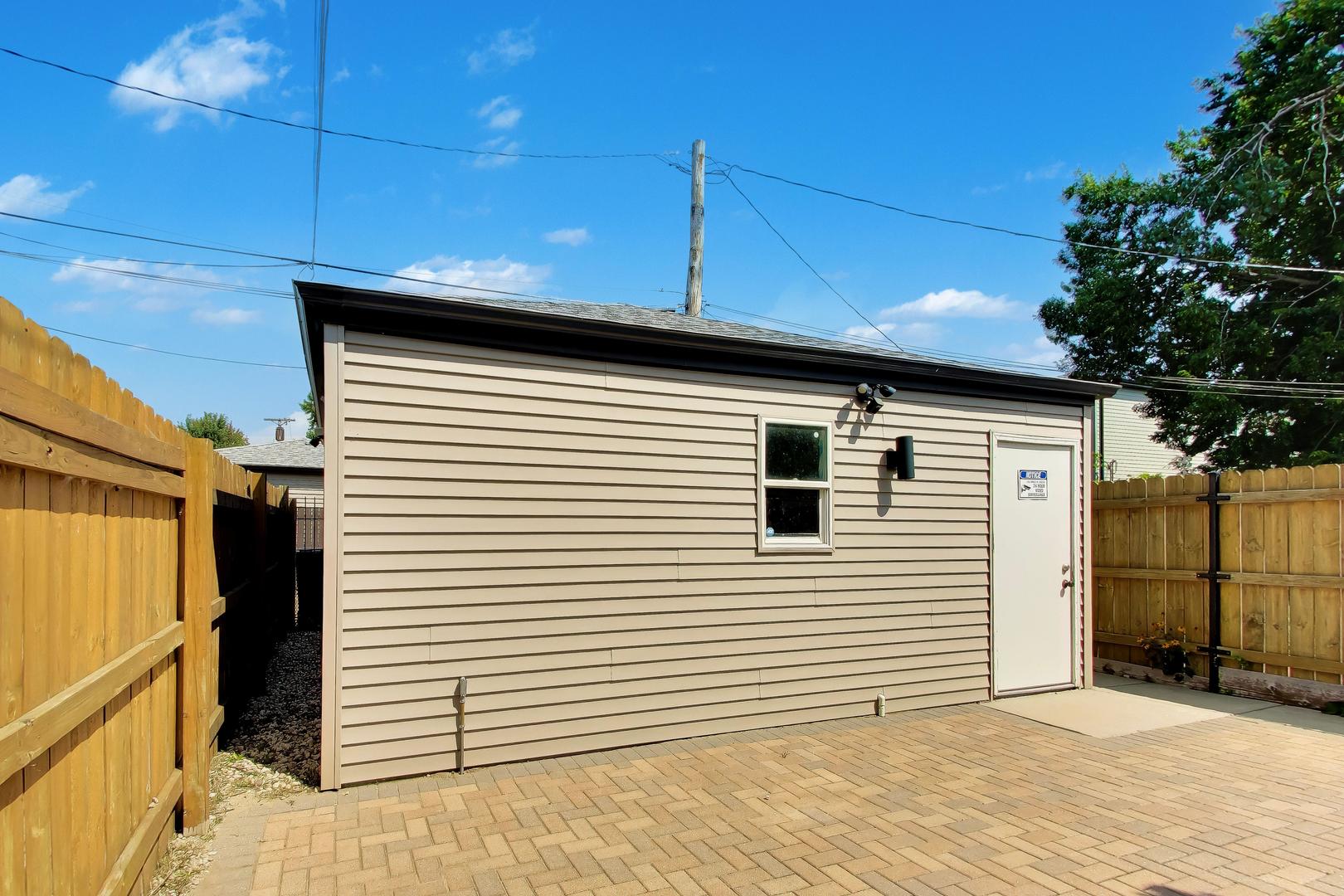 Beige-sided outbuilding with a white door, enclosed by wood fencing beside a brick-paver patio.