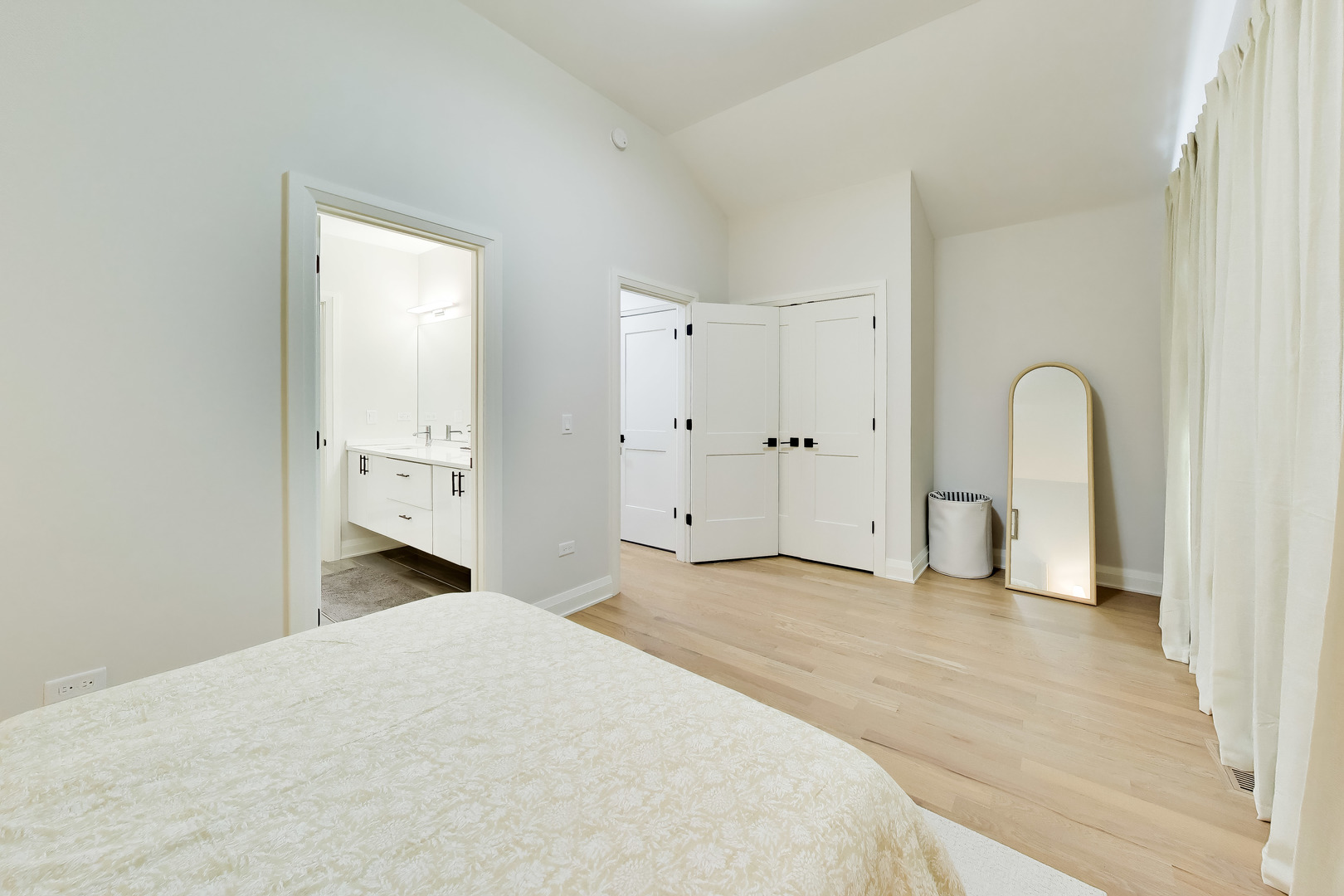 Staged bedroom with a bed, arched floor mirror, and open bathroom doorway.