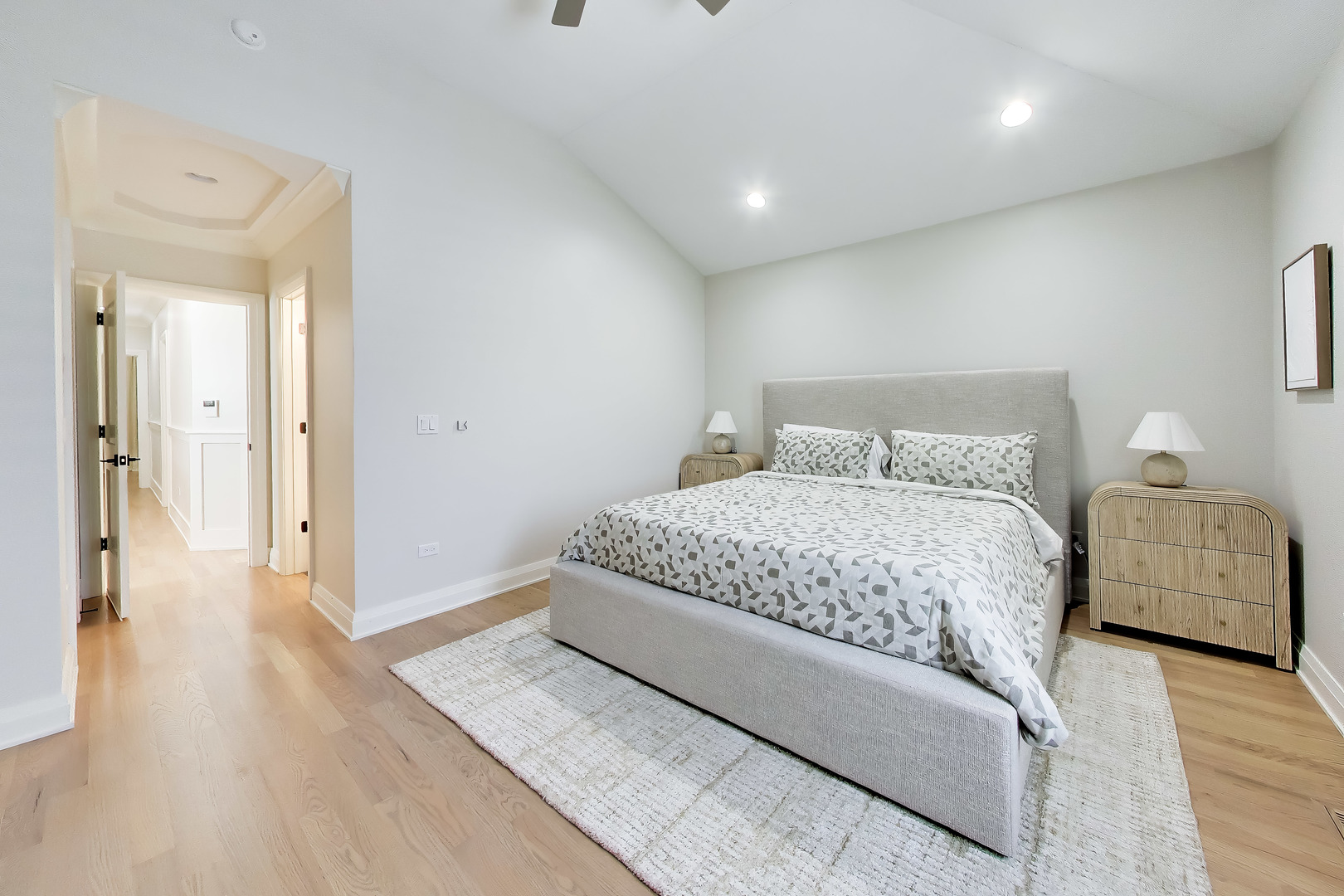 Staged bedroom with patterned bedding, light wood flooring, and an open hallway.