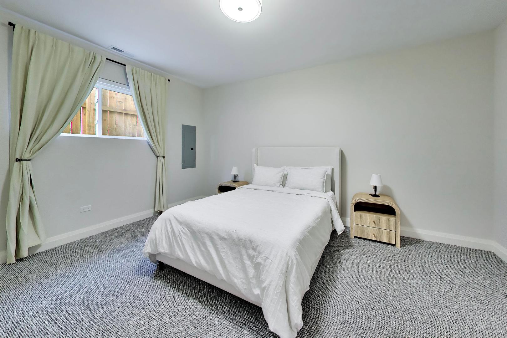 Furnished bedroom with a white bed, gray carpet, and curtains framing the window.