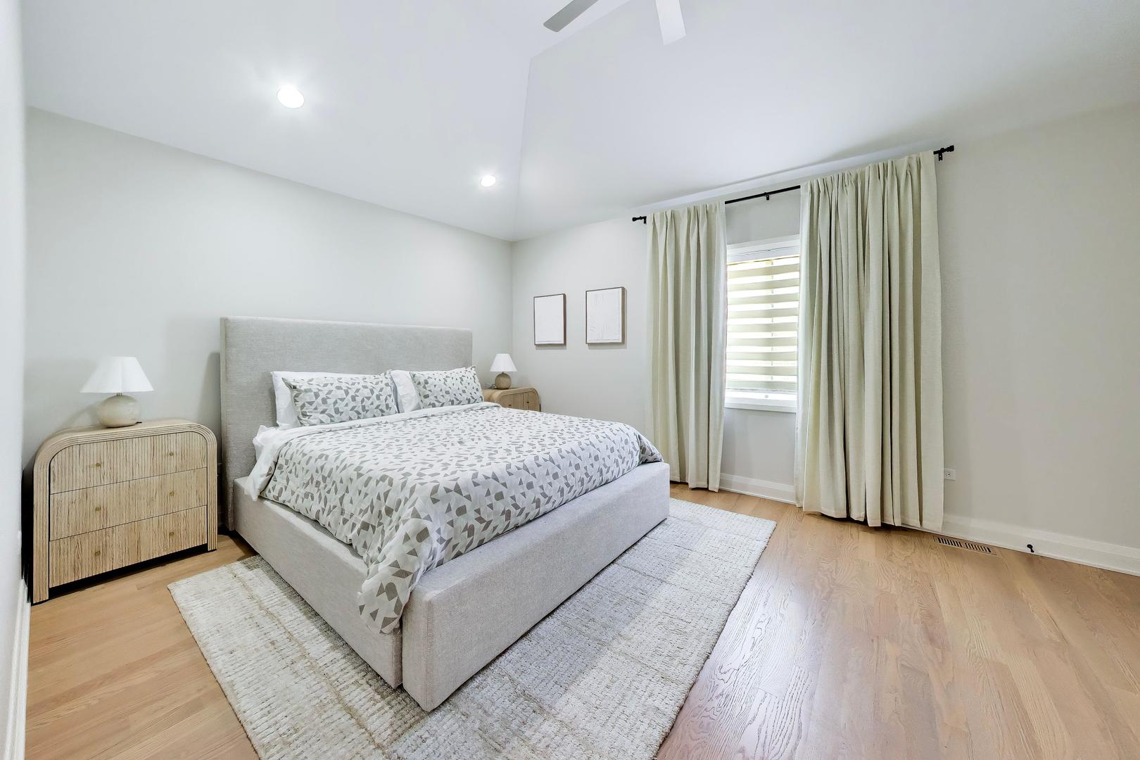 Staged bedroom with a gray upholstered bed, patterned bedding, and cream curtains over a window.