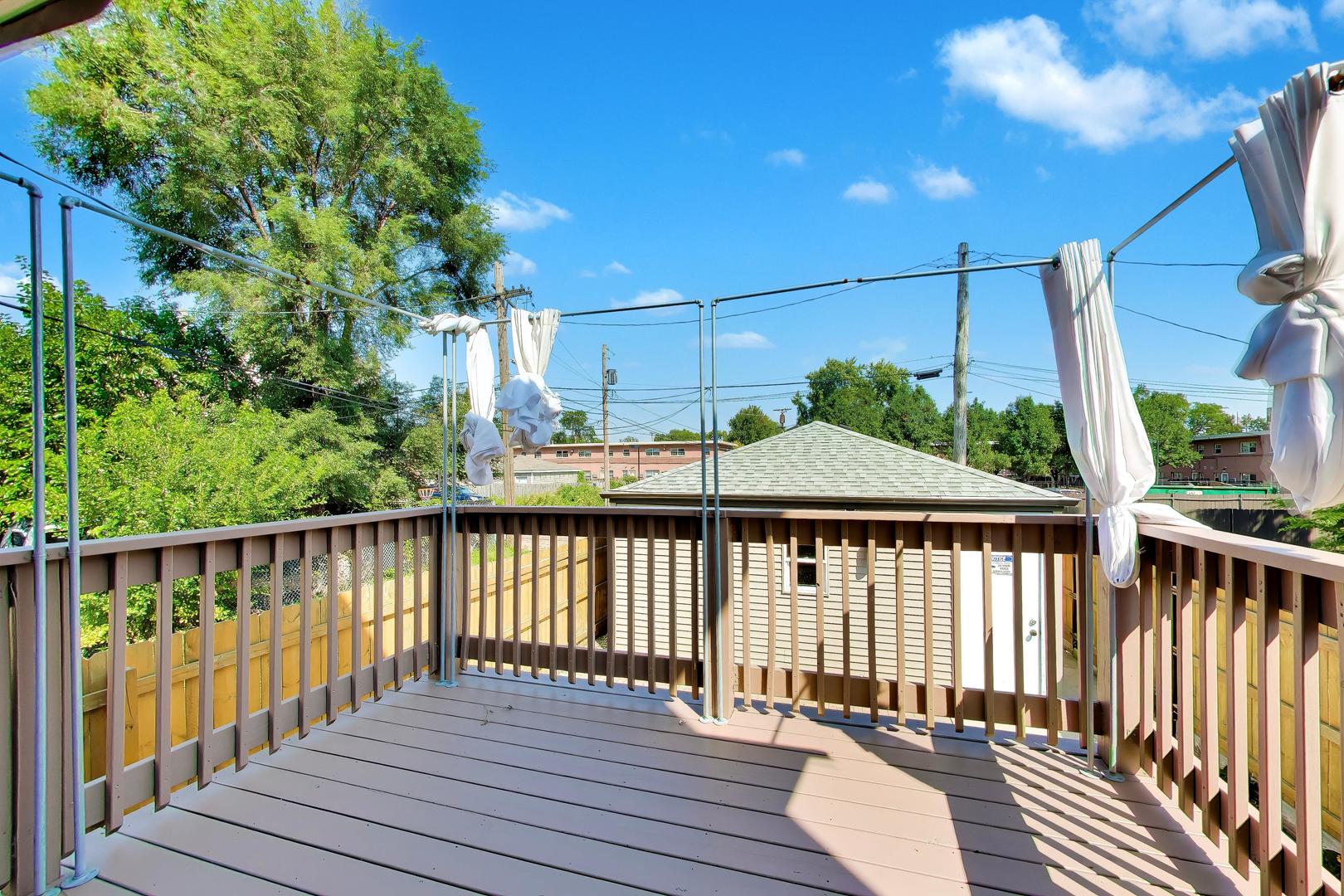 Elevated wooden deck with tied white curtains, leafy trees, and neighboring homes beyond the railings.