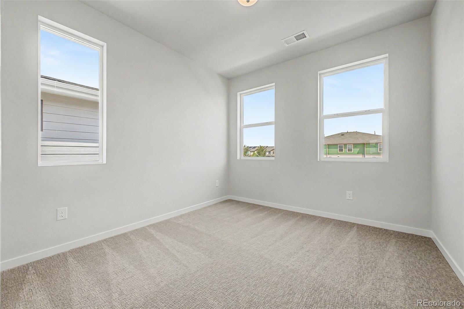 Vacant carpeted room with white walls and three windows overlooking neighboring homes.