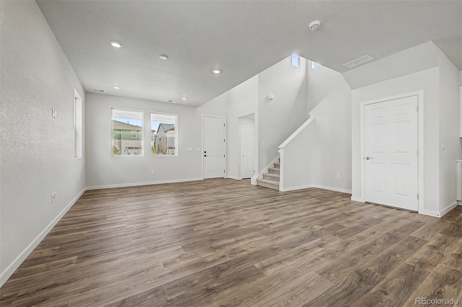 Vacant living room with wood-look flooring, two windows, and a staircase beside the entry doors.