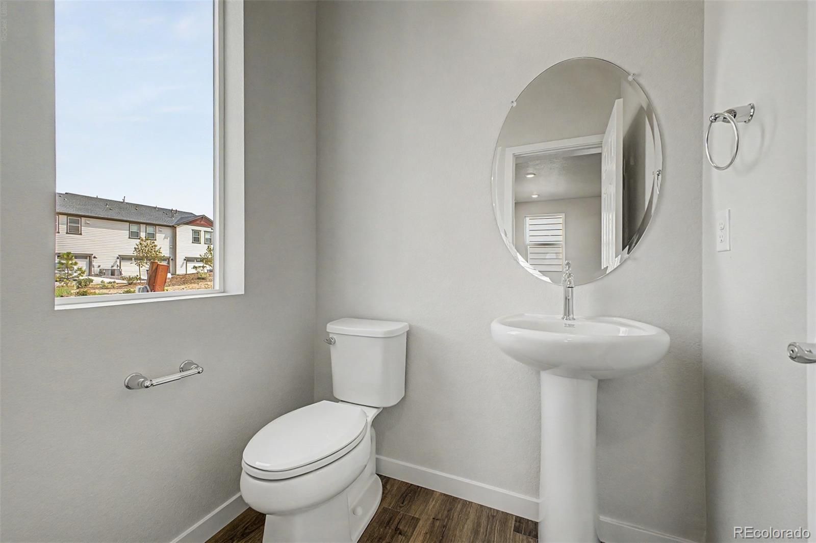 Bathroom with a pedestal sink and oval mirror beside a window overlooking neighboring homes.