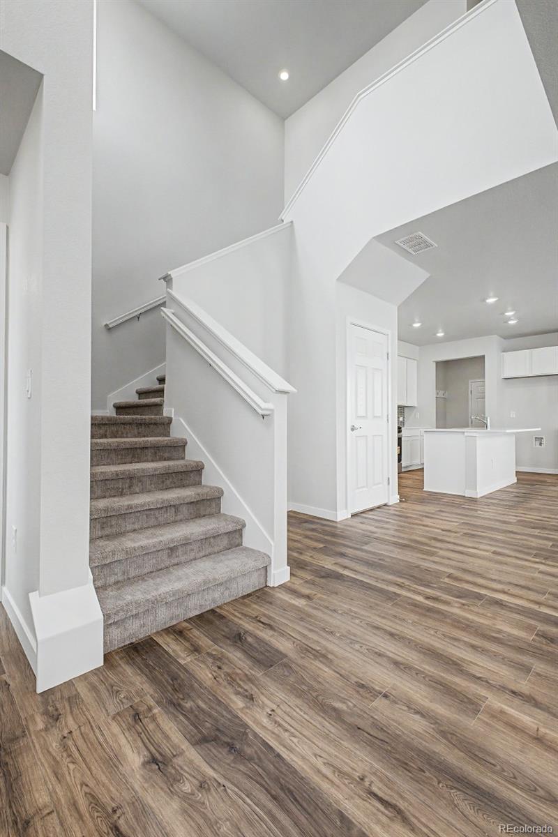 Carpeted staircase beside an open kitchen, with tall ceilings and wood-look flooring.