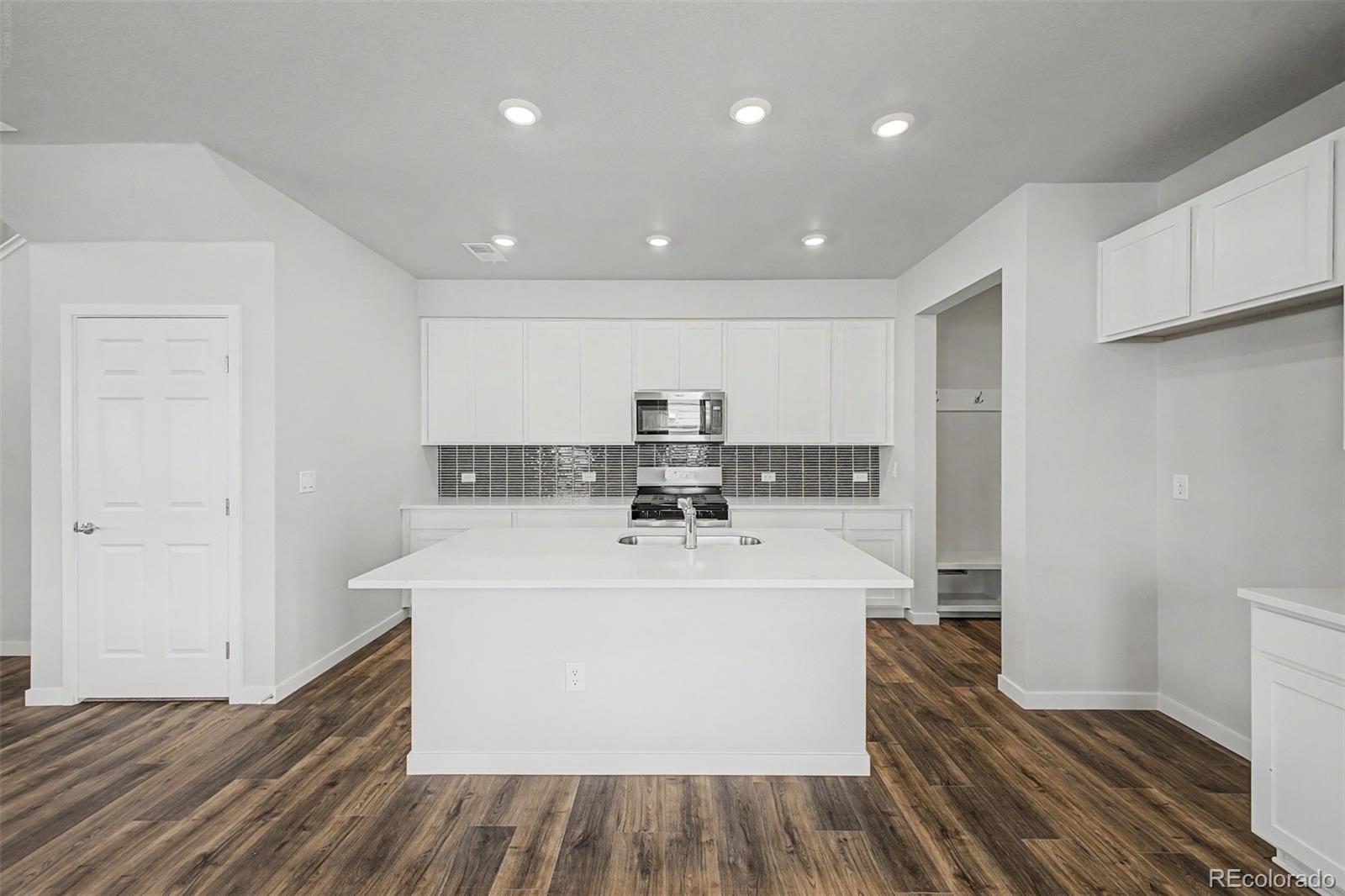 Vacant kitchen with a large white island, dark tile backsplash, and wood-look flooring.