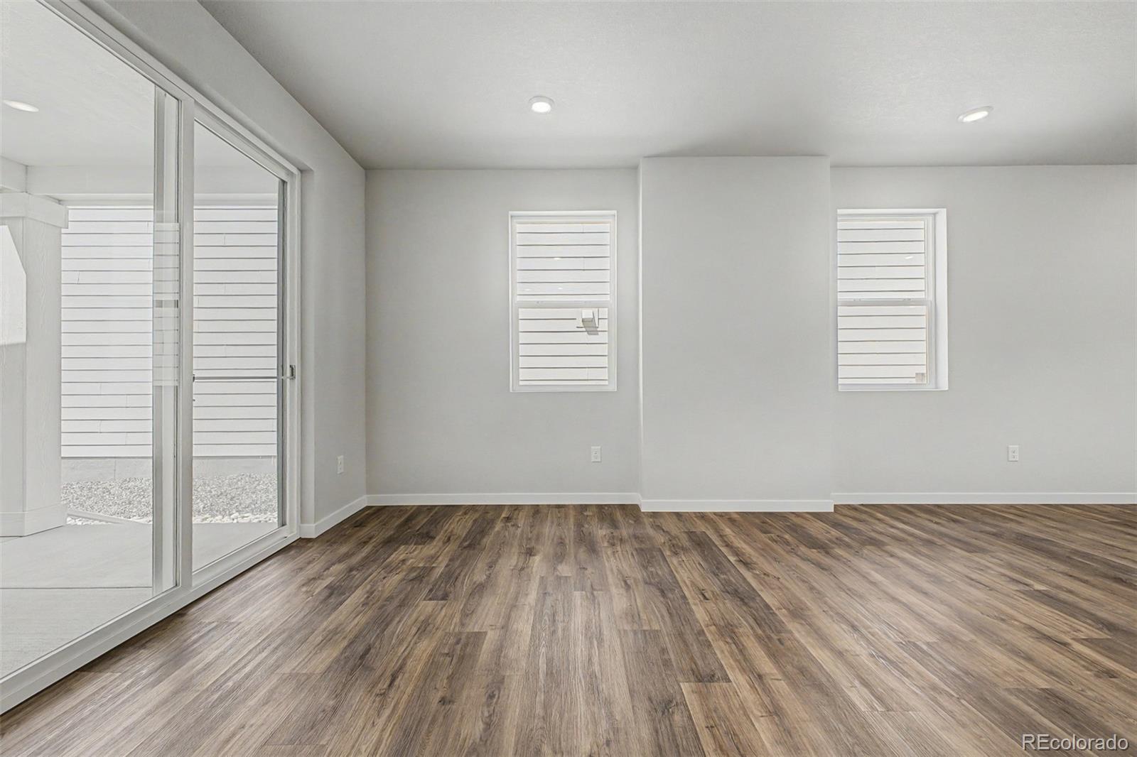 Vacant room with wood-look flooring, two windows, and a sliding glass door to a gravel patio.