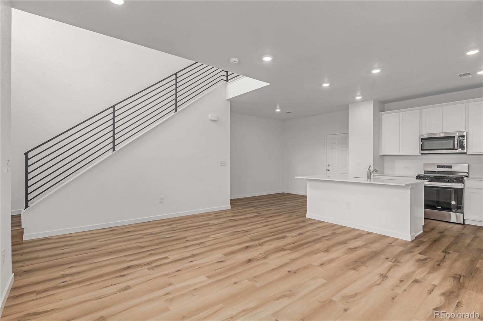 Open kitchen and living area with a white island, stainless appliances, and a staircase with black railing.