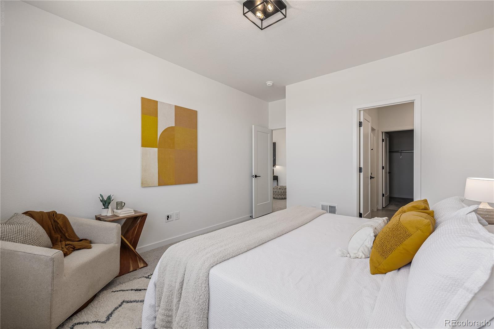 Staged bedroom with a white bed, mustard pillows, and a beige armchair beneath geometric wall art.