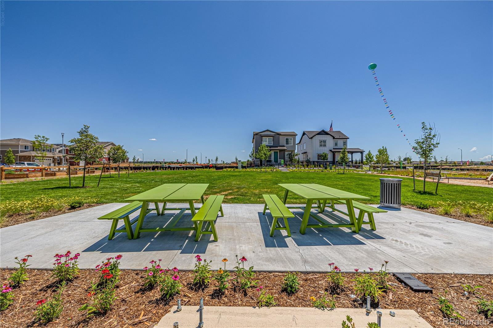Community park patio with green picnic tables, flower beds, and homes in the background.