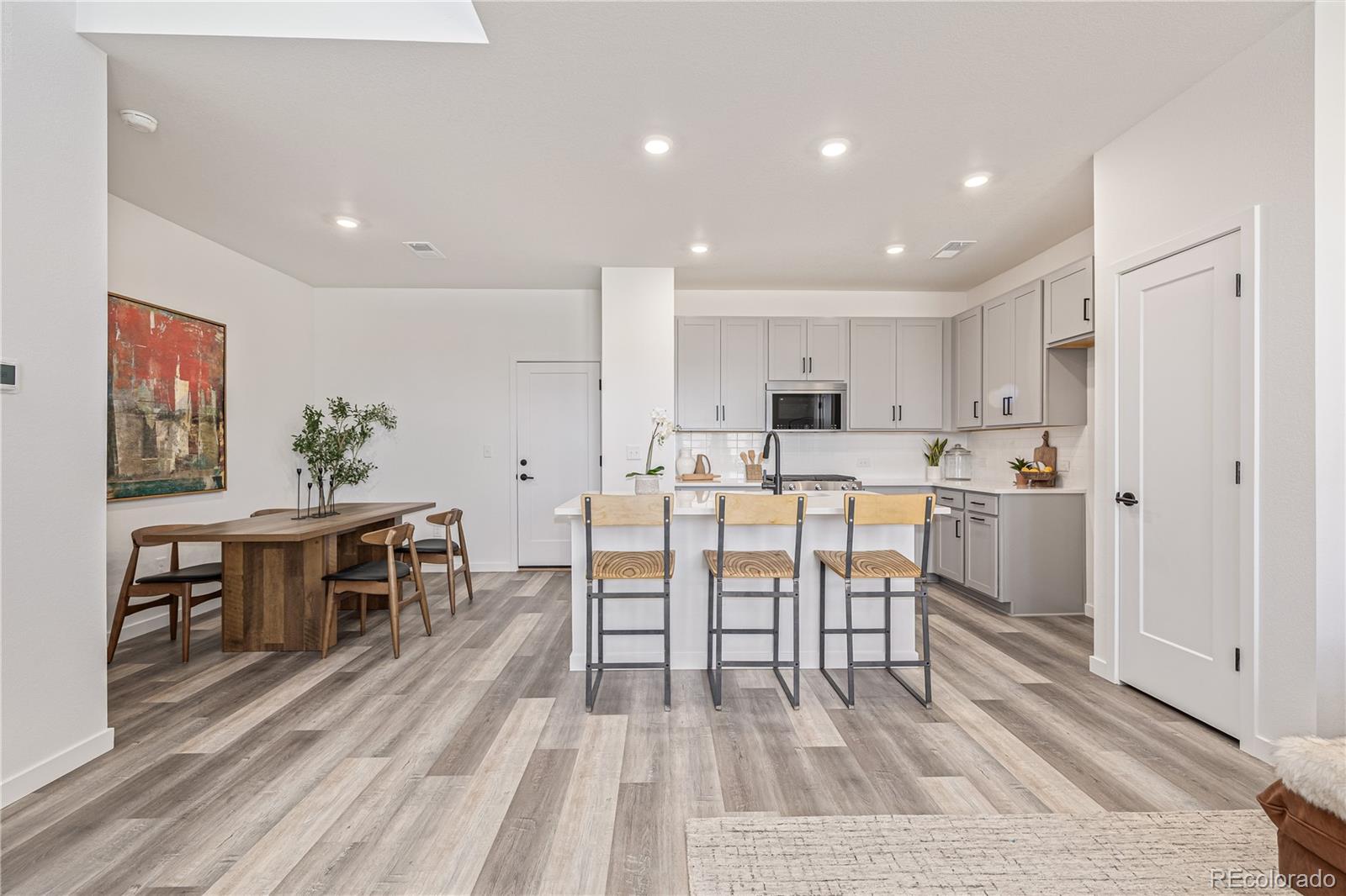 Open kitchen and dining area with a white island, three bar stools, and gray plank flooring.