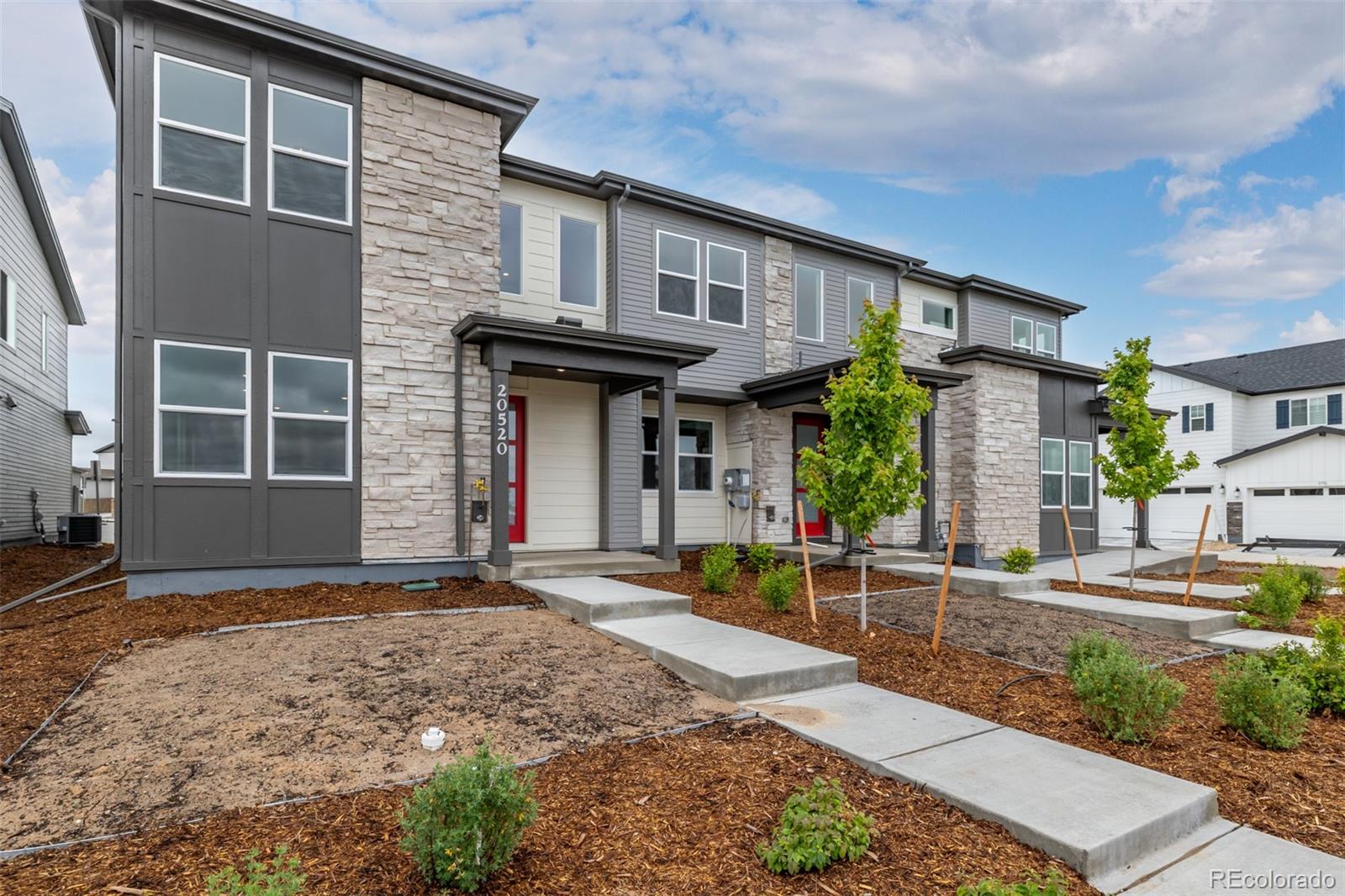 Modern townhome with gray siding, stone accents, red doors, and newly planted landscaping.