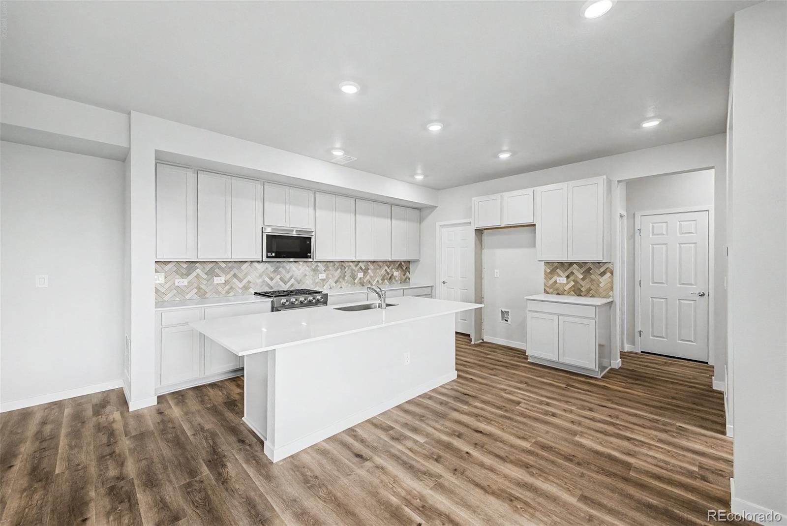Vacant kitchen with a large white island, herringbone backsplash, and wood-look flooring.