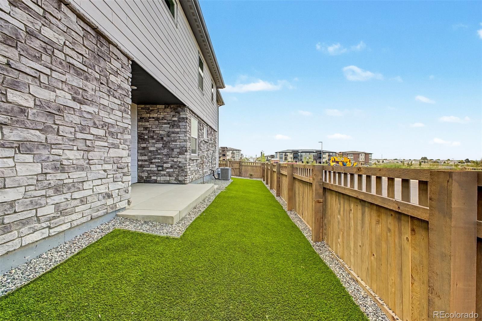 Side yard with artificial turf, a small concrete patio, and a wooden fence beside a stone-sided house.