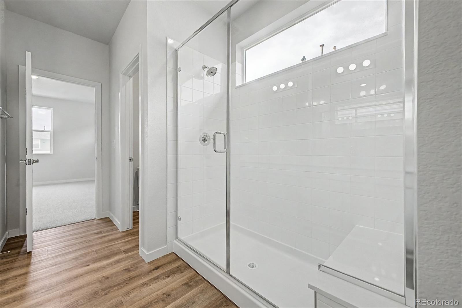 Bathroom with a glass-enclosed white-tiled shower, high window, and wood-look flooring.
