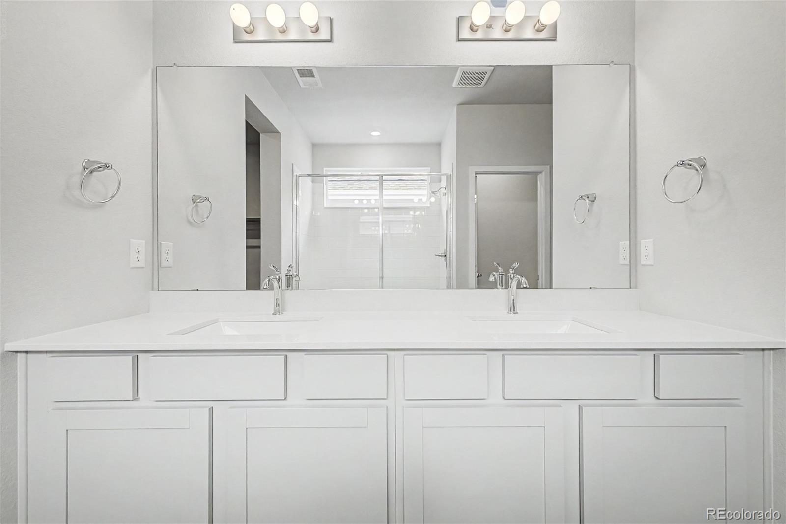 Bright bathroom with a white double-sink vanity and glass shower reflected in a large mirror.