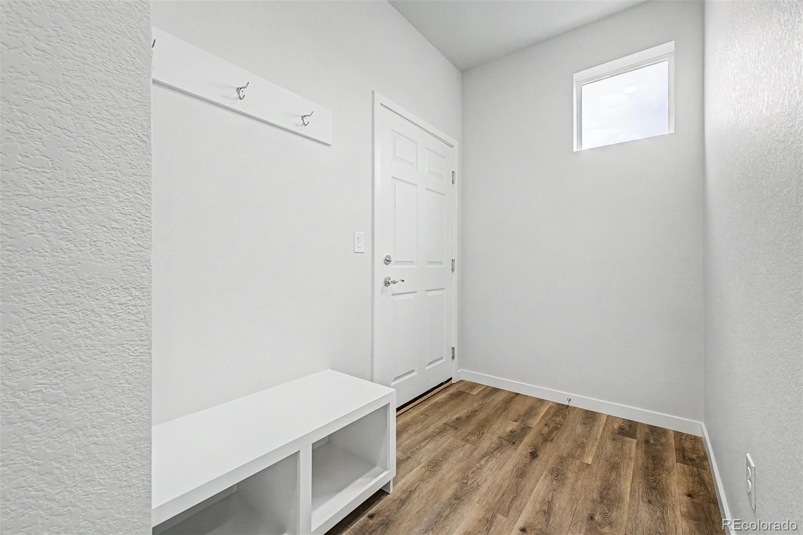 Bright entryway with a high ceiling, wall hooks, white bench, and wood-look flooring.