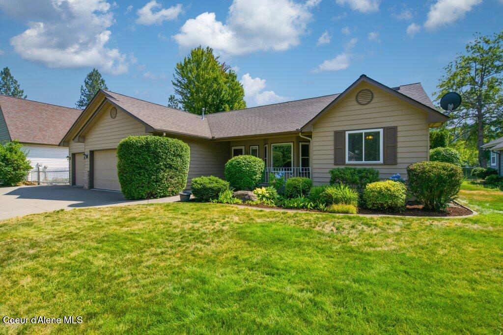 Single-story beige home with an attached garage, covered entry, landscaped shrubs, and a broad green lawn.