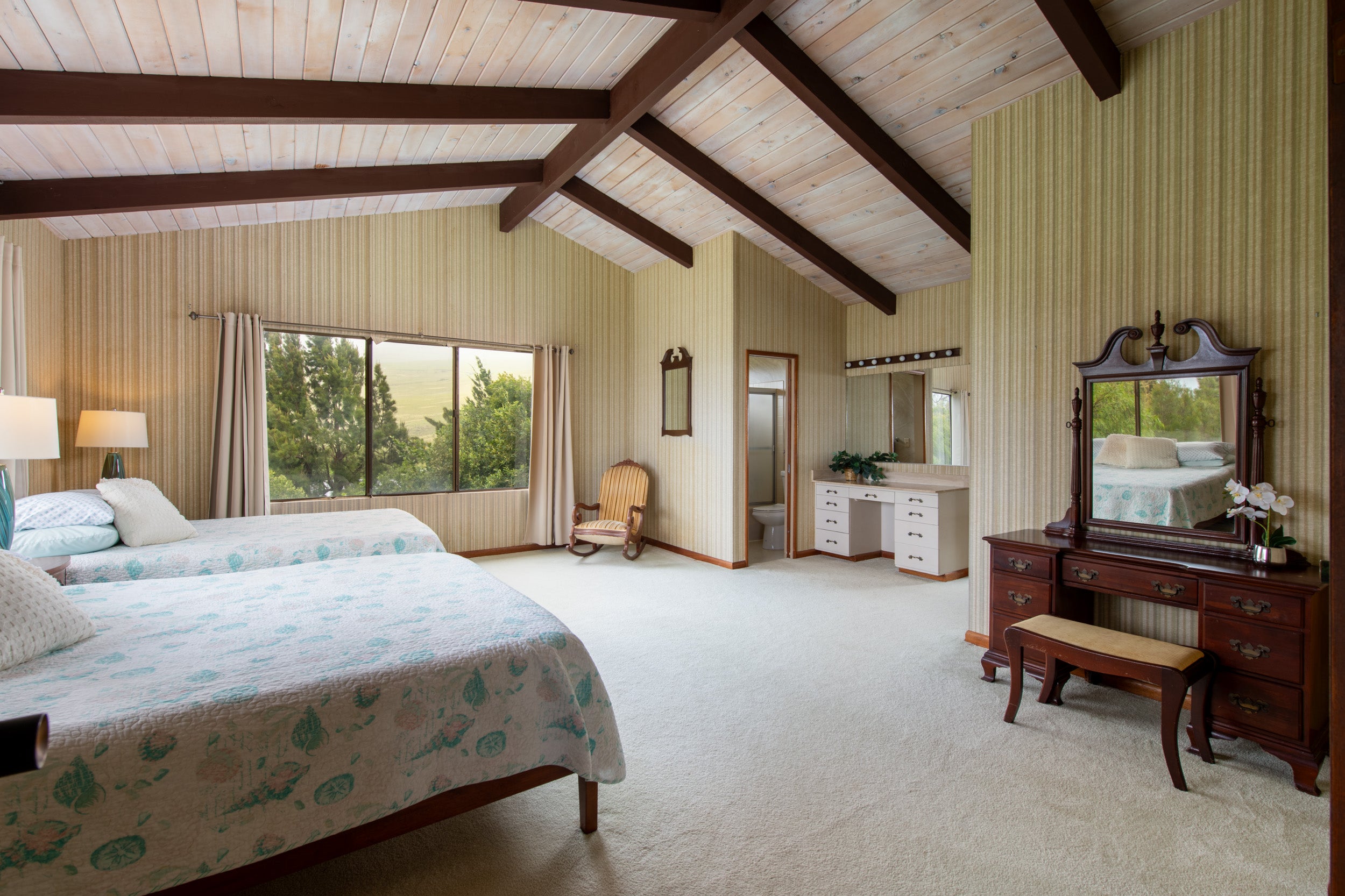 Bedroom with two beds, exposed ceiling beams, a vanity, and a window overlooking trees and open land.
