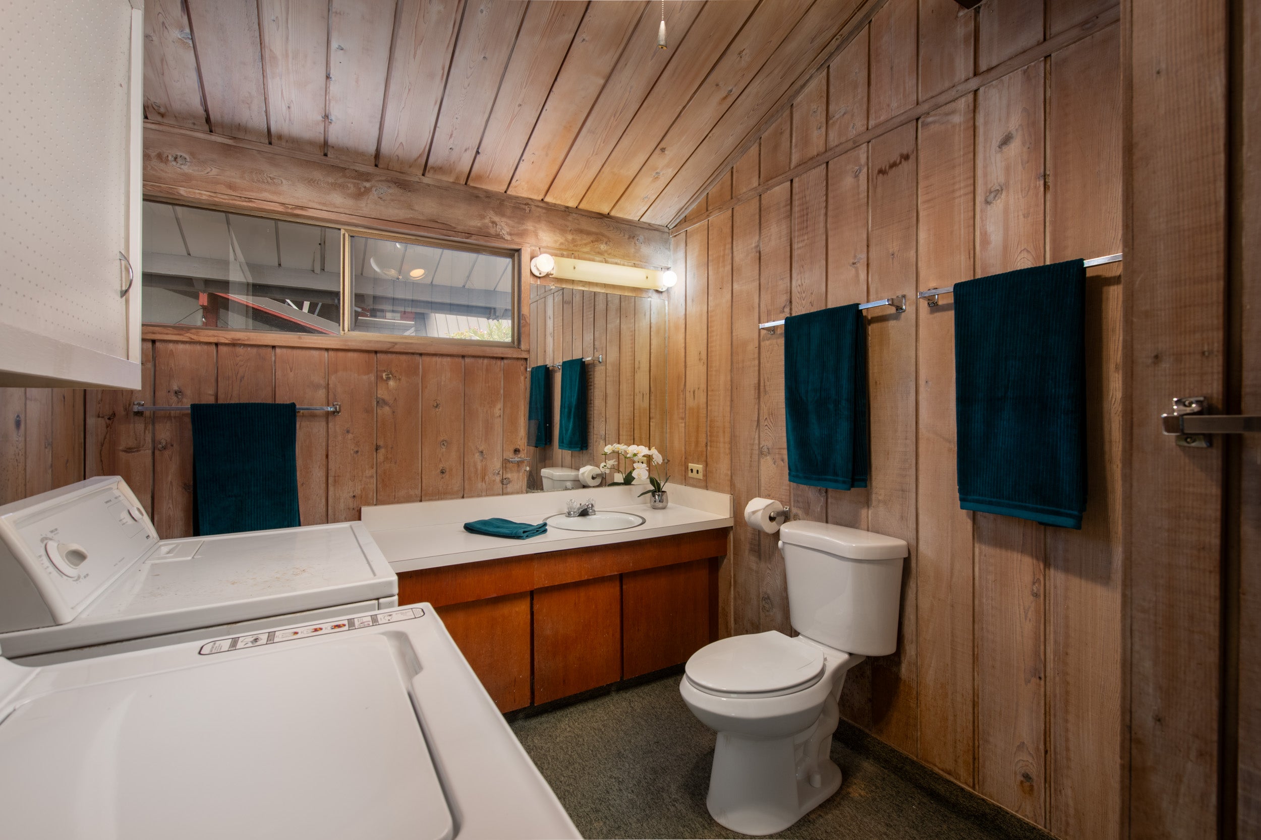 Wood-paneled bathroom with washer and dryer, white vanity, toilet, and teal towels.