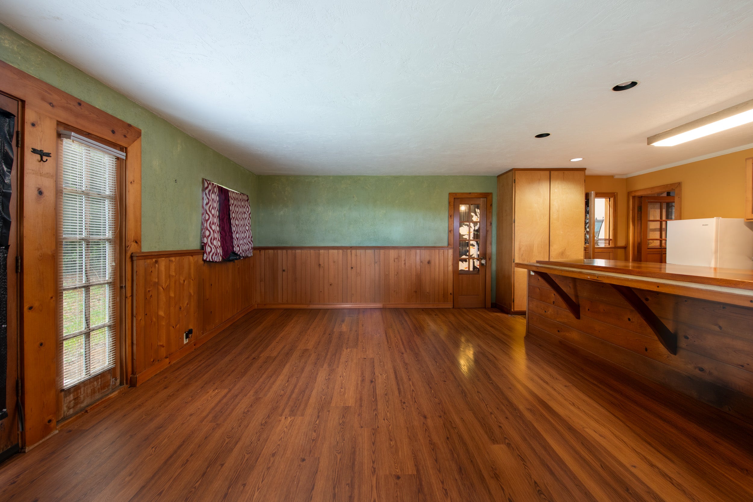 Mostly empty kitchen with wood floors, paneled walls, and a long counter beside a white refrigerator.