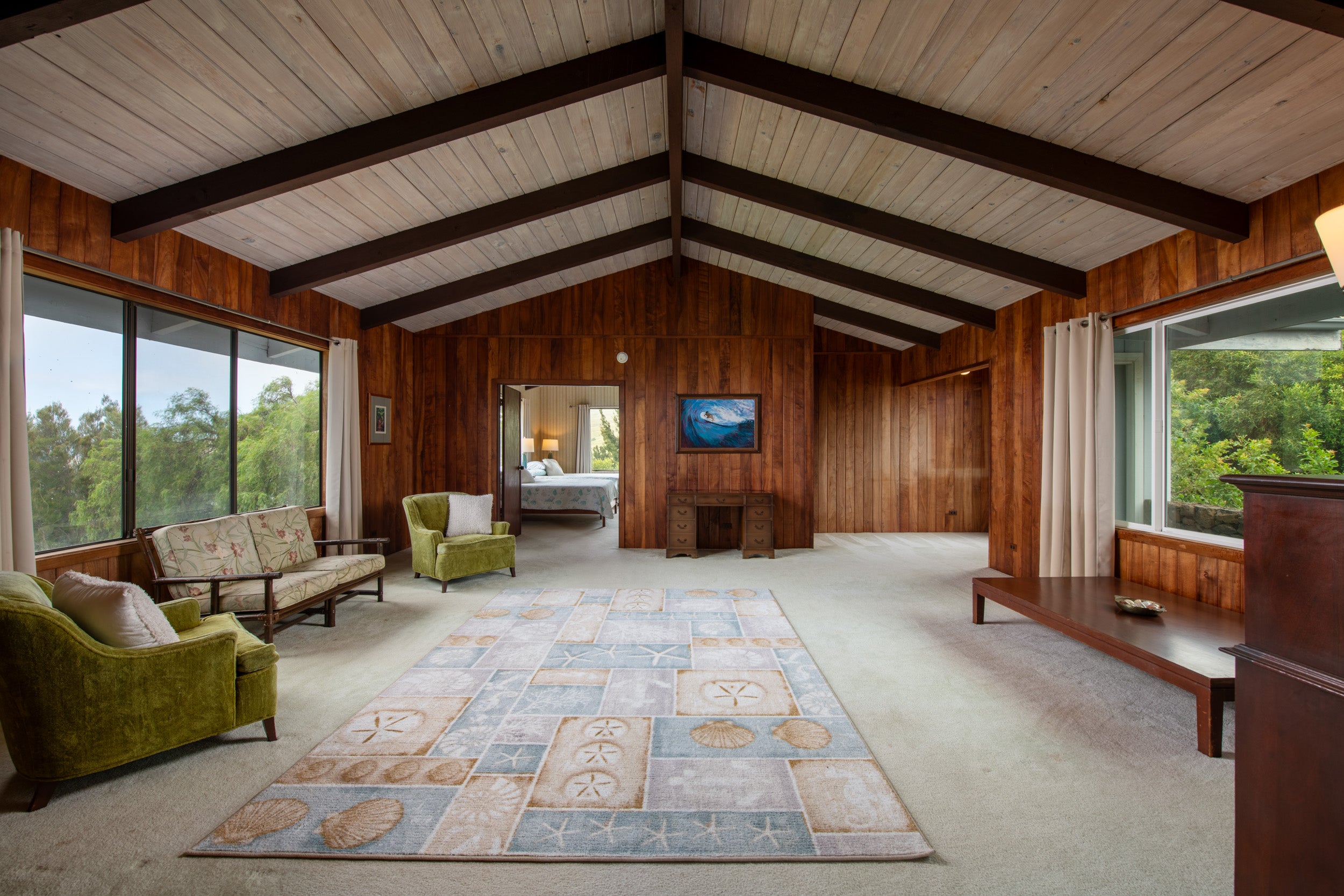 Wood-paneled living room with vaulted beam ceiling, large windows, and a patterned rug.