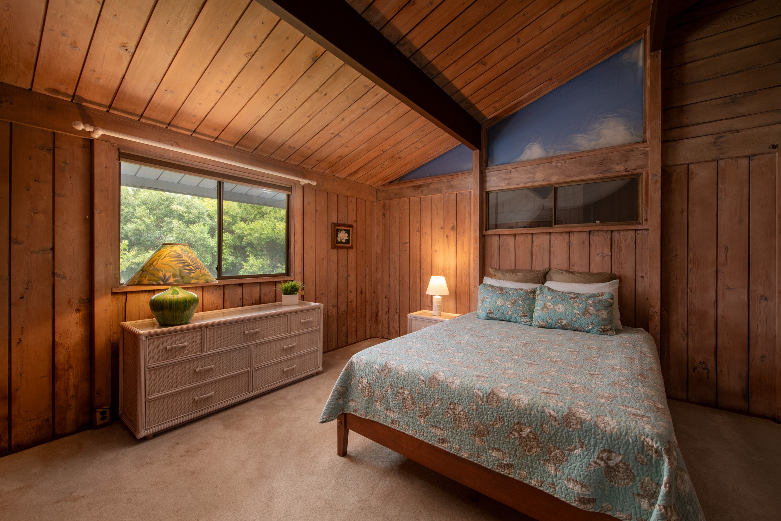 Furnished bedroom with a blue floral quilt, wood-paneled walls, and a high sloped ceiling.