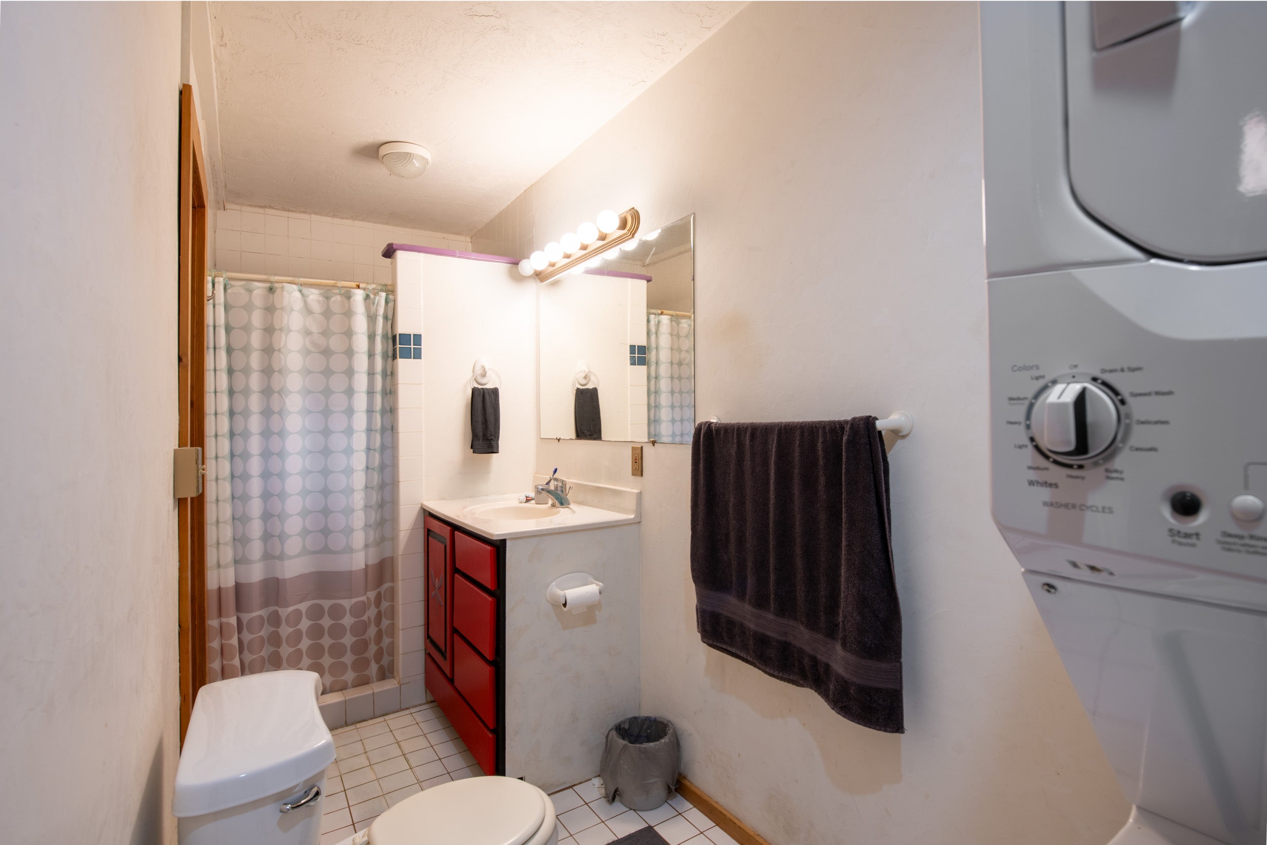 Compact bathroom with red vanity, patterned shower curtain, and washer and dryer at the right edge.
