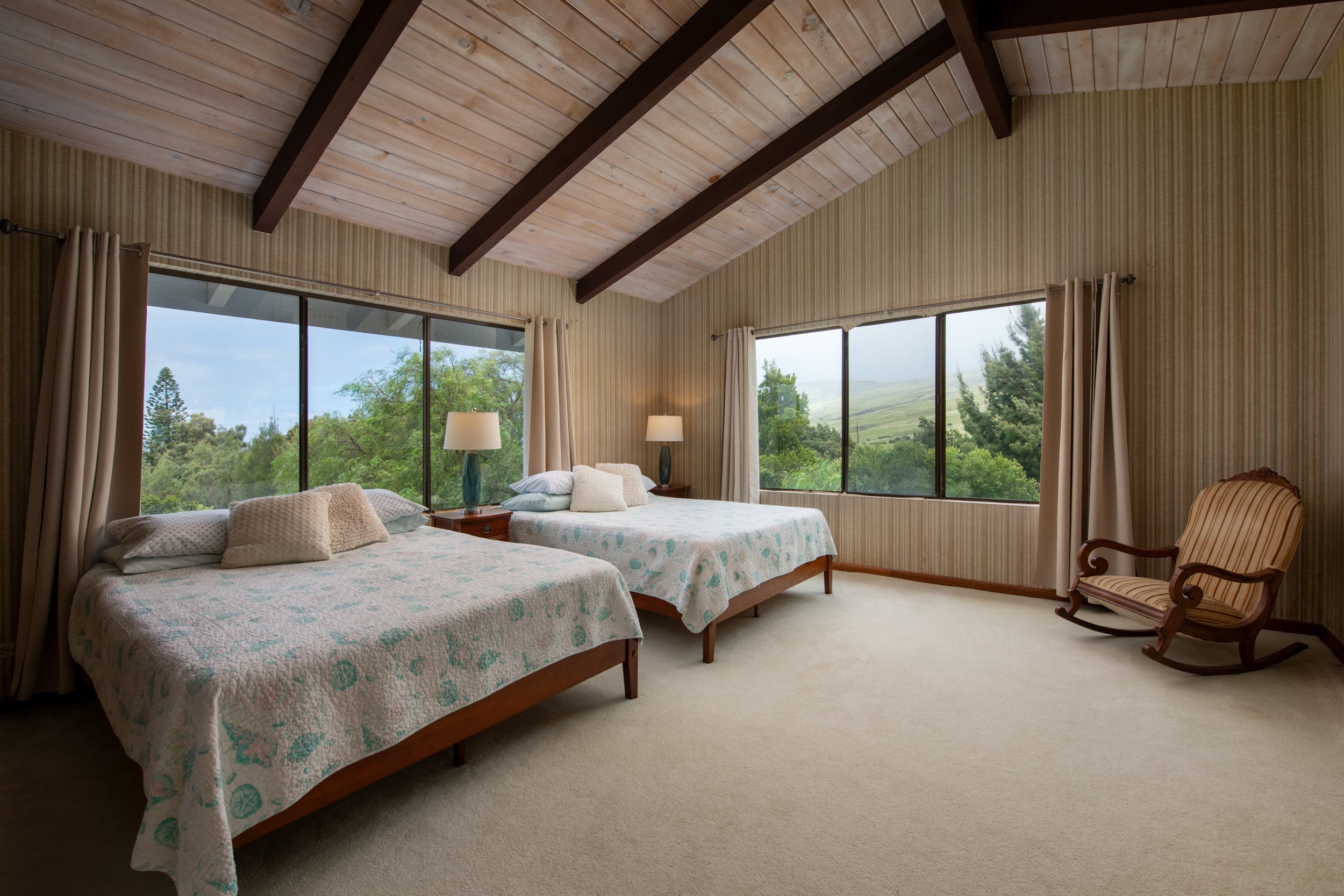 Bedroom with two beds beneath a vaulted wood ceiling, large windows, and a wooden rocking chair.