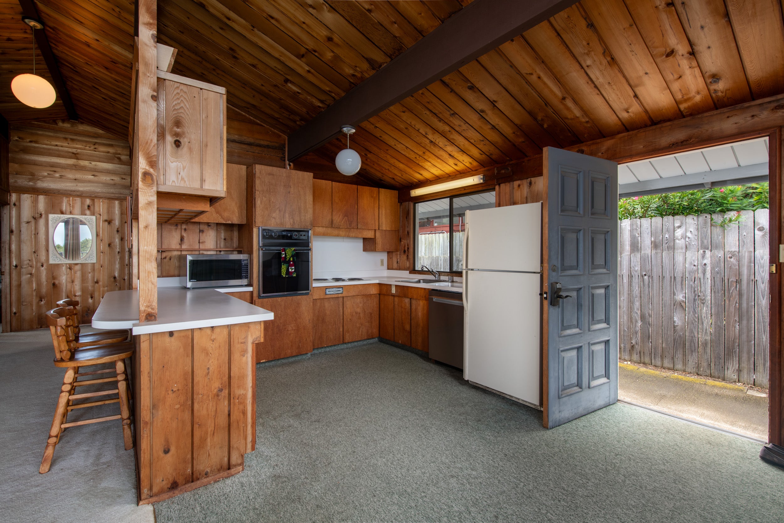 Wood-paneled kitchen with a high timber ceiling, breakfast counter, and open door to a fenced yard.
