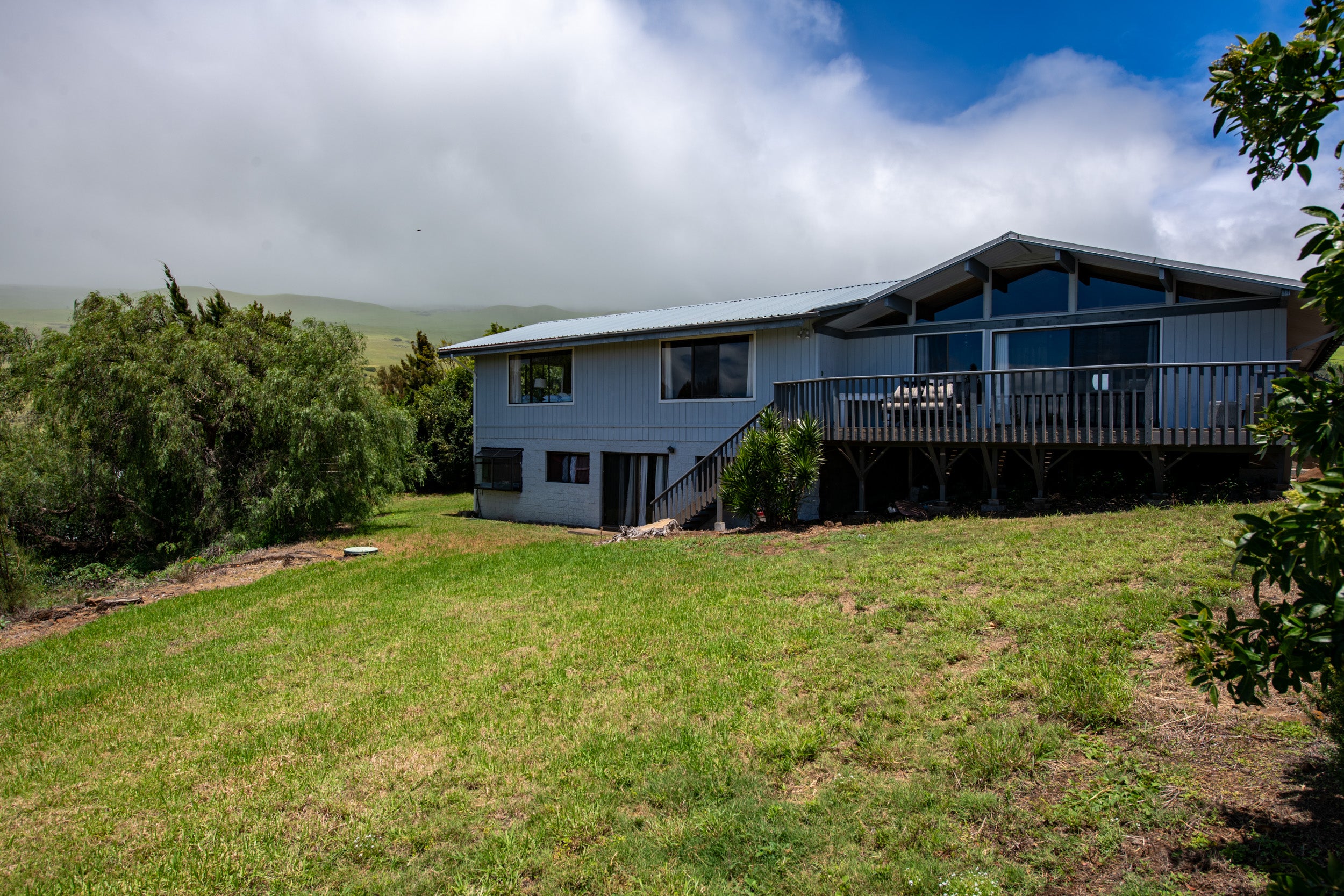 Gray raised home with a broad deck overlooking a sloping lawn and green hills.