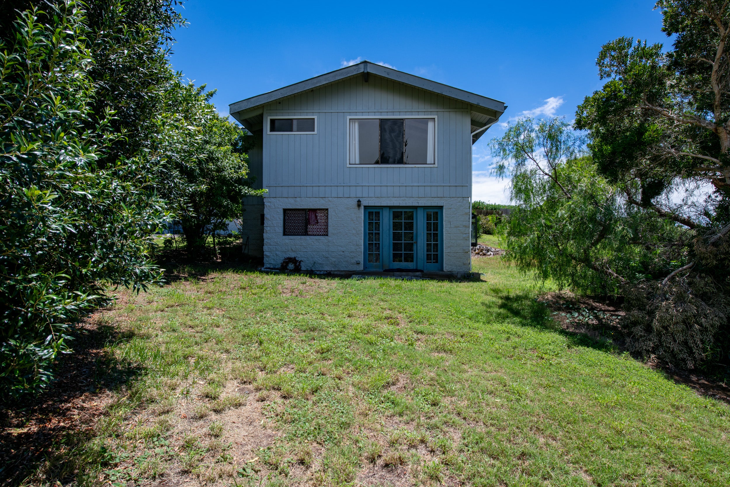 Two-story blue house with teal French doors and a grassy yard framed by mature trees.