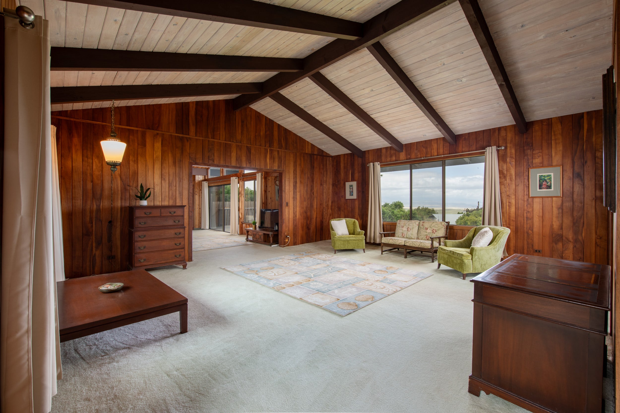 Living room with vaulted wood-beamed ceiling, wood-paneled walls, green chairs, and an ocean view through the window.