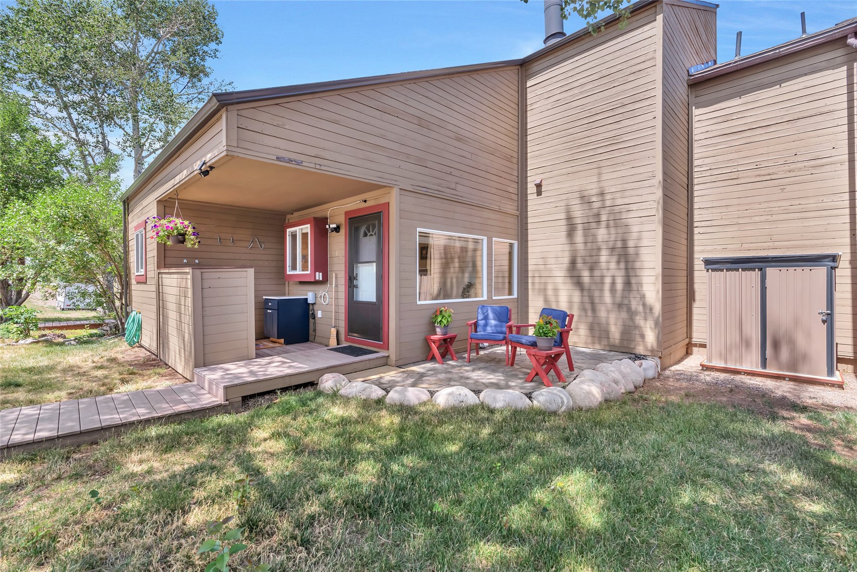 Wood-sided home with a covered entrance, deck, and stone-edged patio with blue chairs.