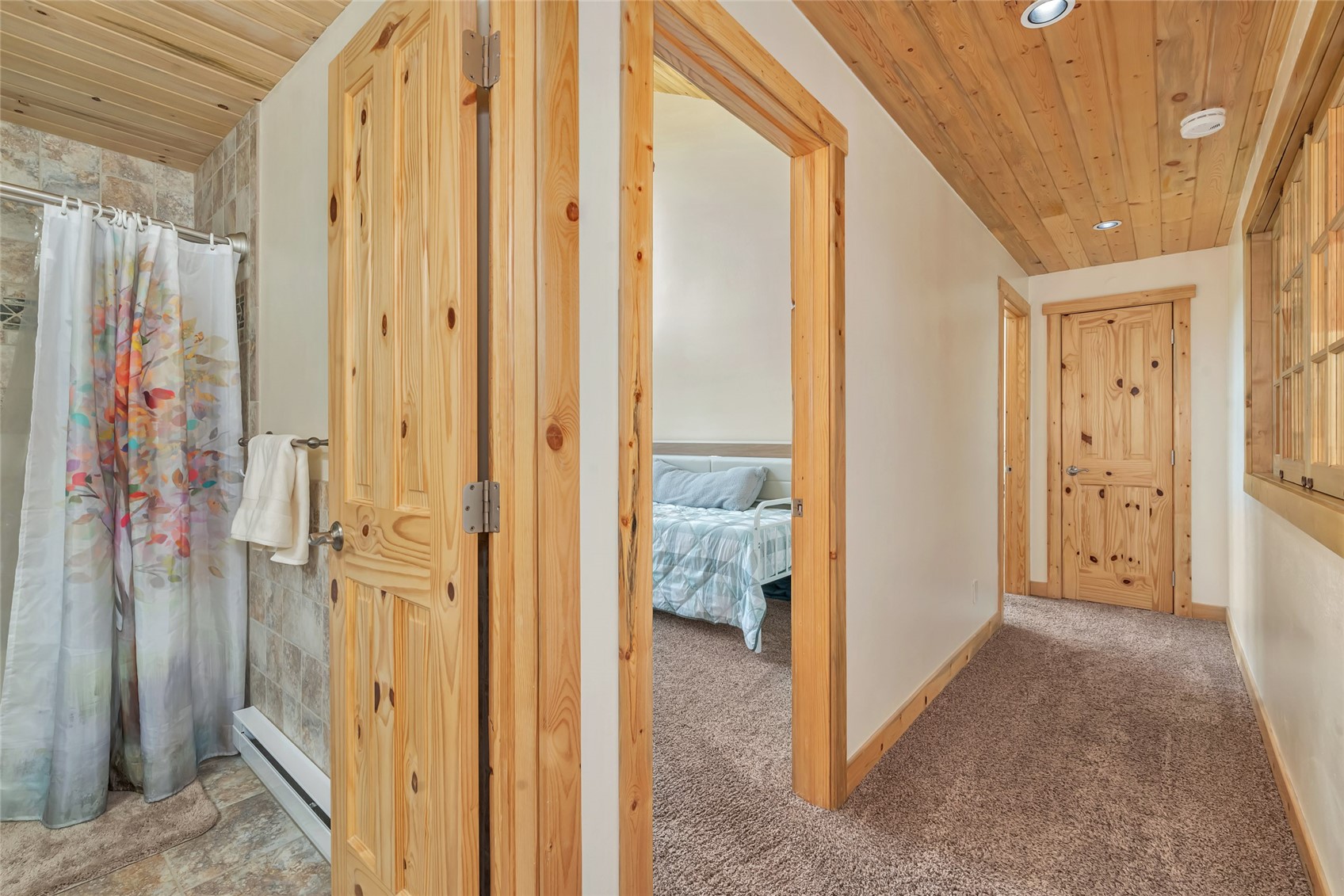 Carpeted hallway with pine doors and a wood ceiling, opening to a bedroom beside a tiled bathroom.