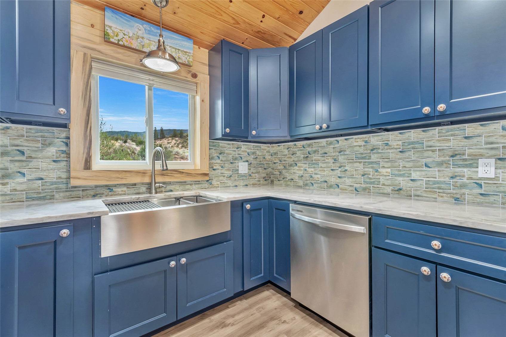 Kitchen with blue cabinets, a stainless farmhouse sink, patterned tile backsplash, and a window overlooking open terrain.