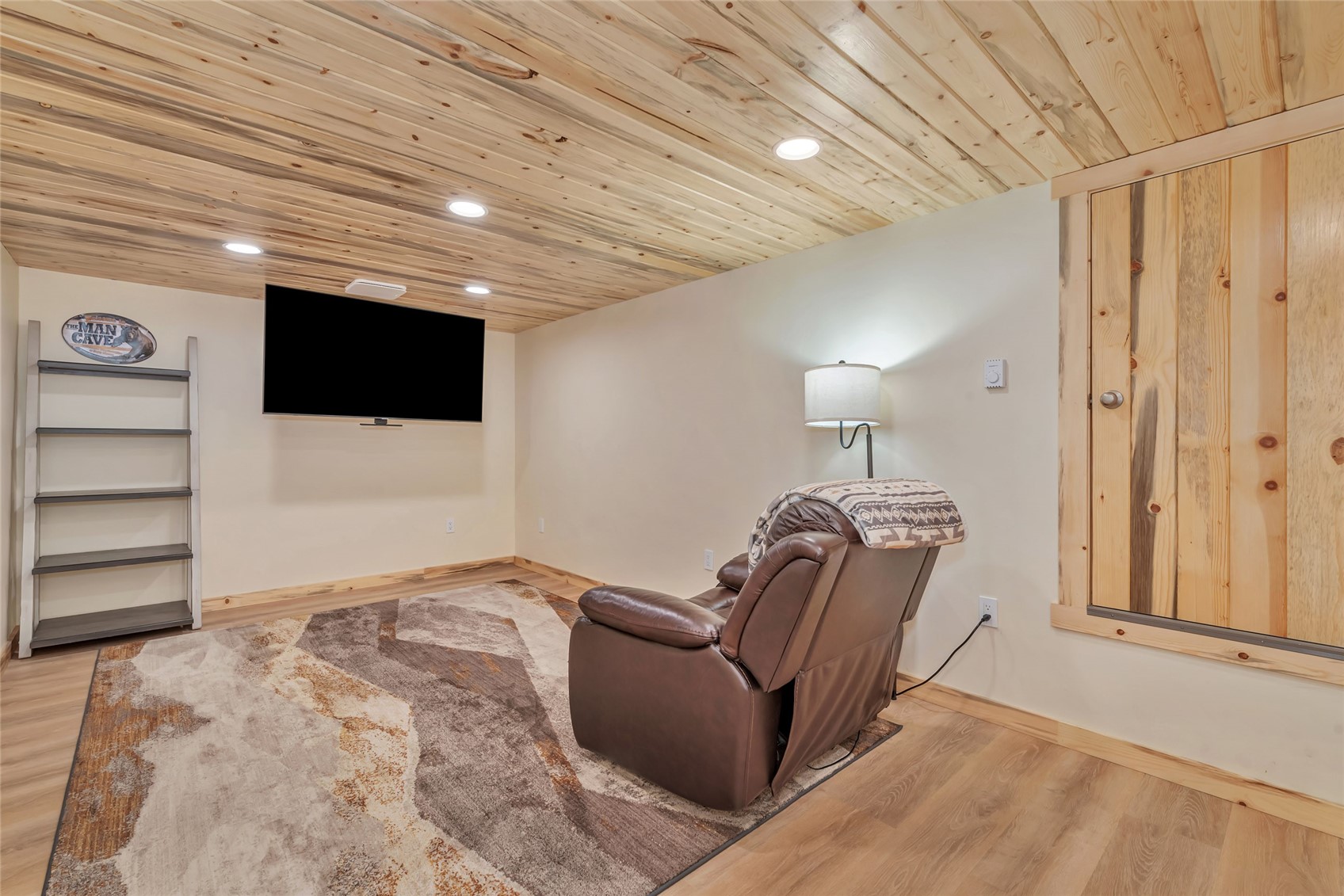 Furnished media room with a leather recliner, wall-mounted TV, and wood-plank ceiling.