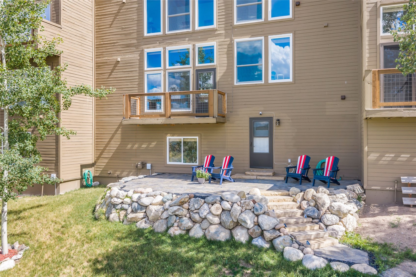 Tan home exterior with a stone retaining wall, patio, and four striped Adirondack chairs.